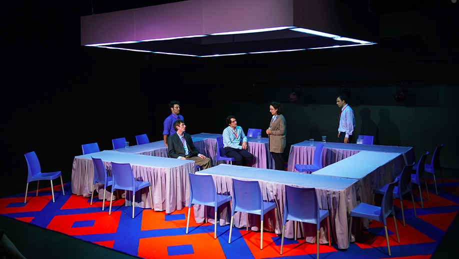 people stand on blue and red stage among white rectangular tables formed into a large rectangle